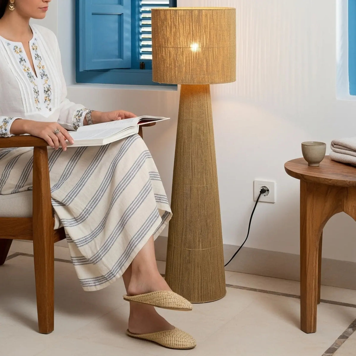 Lampadaire en jute allumé près d'une femme qui lit dans un fauteuil en bois, devant un volet bleu