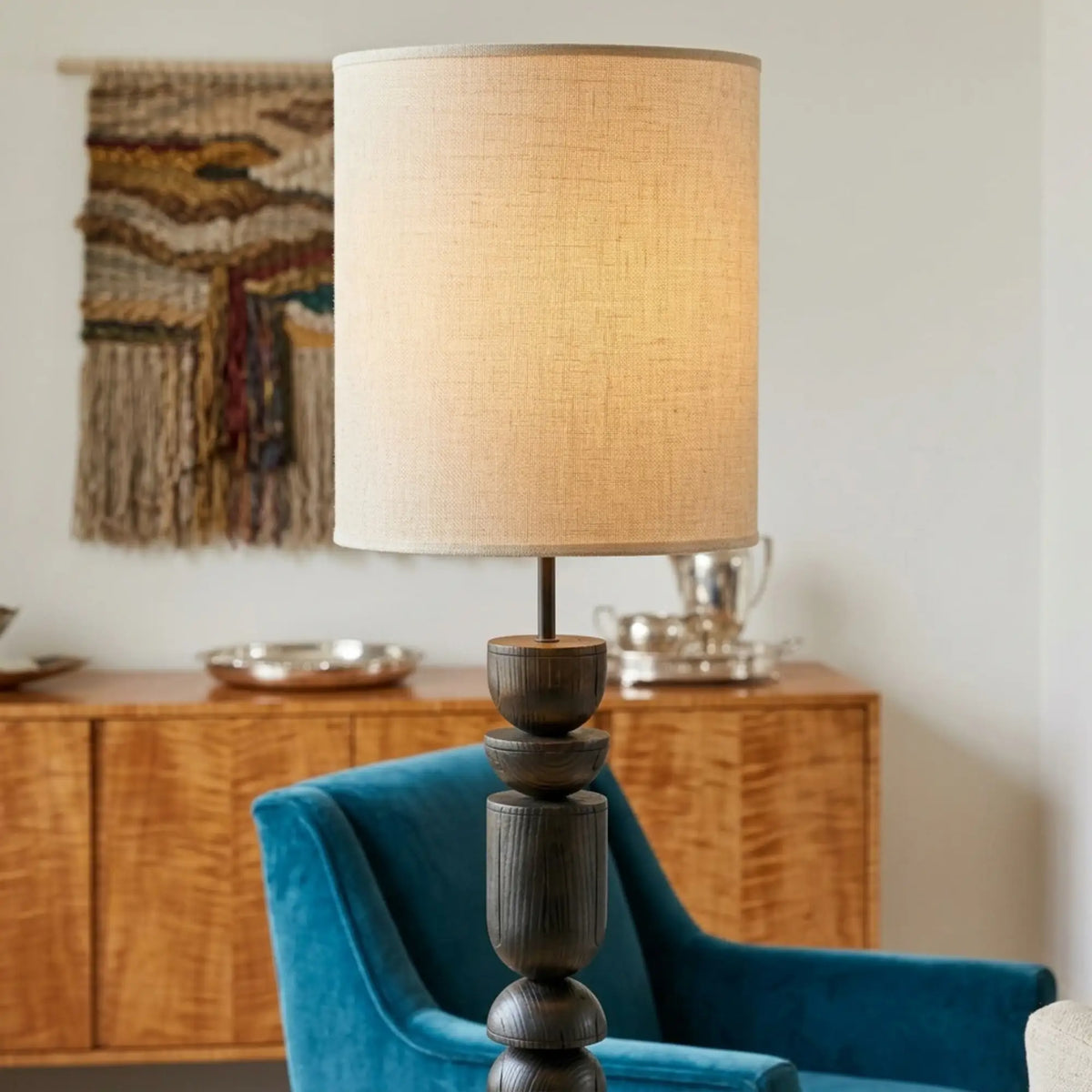 Gros plan sur l'abat-jour cylindrique en lin et le haut du pied en bois tourné, fauteuil en velours bleu canard et buffet en bois