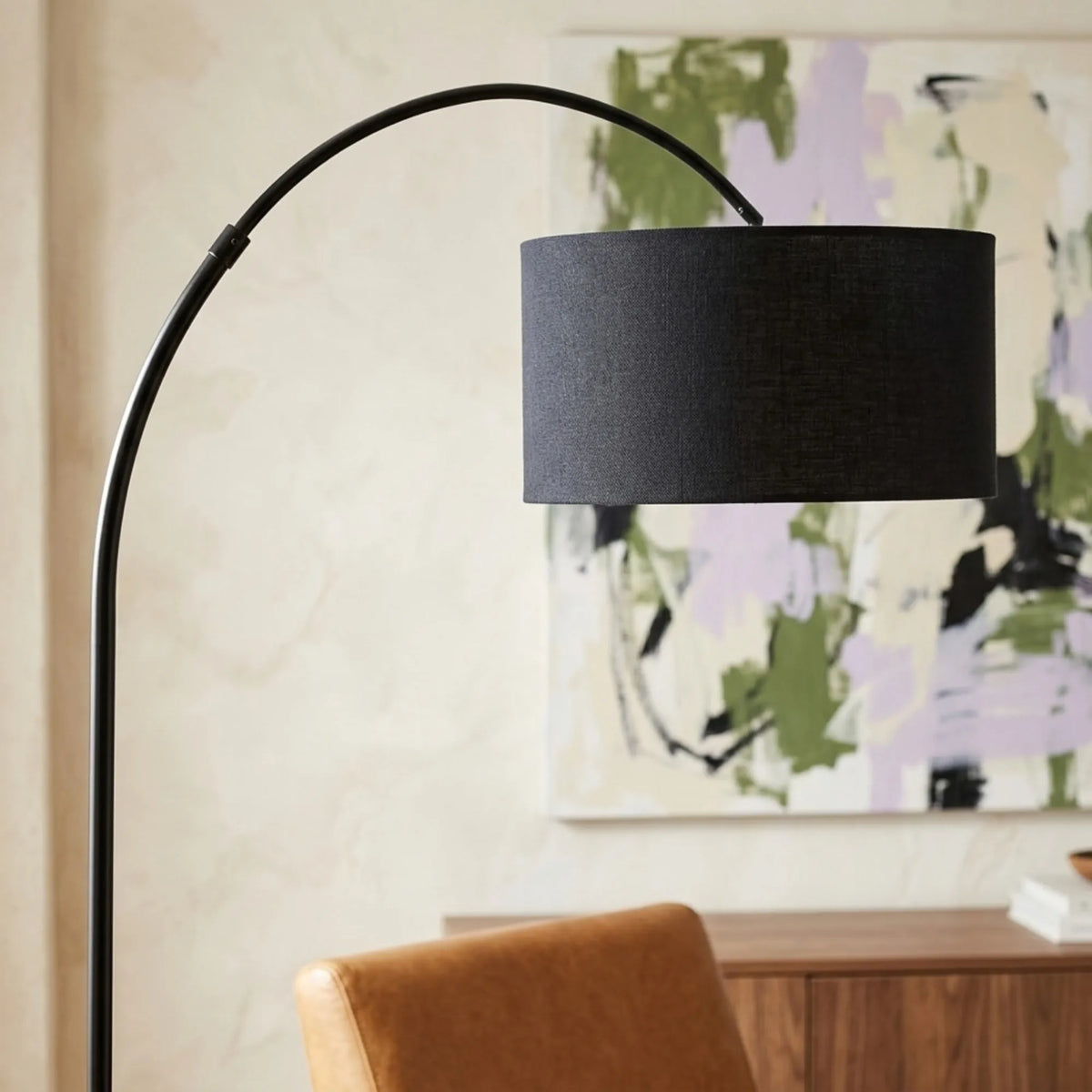 Gros plan sur l'abat-jour tambour en tissu noir et la tige arquée du lampadaire, devant un tableau abstrait vert et lilas