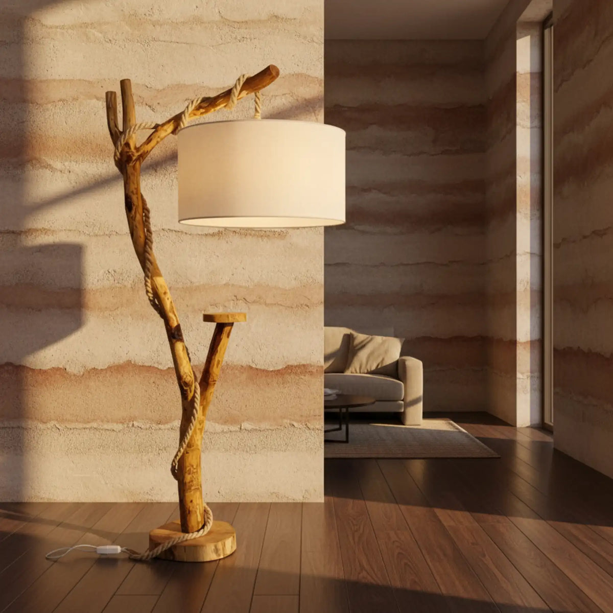 Lampadaire branche en bois avec abat-jour tambour en tissu allumé, devant un mur aux teintes sable et un parquet foncé