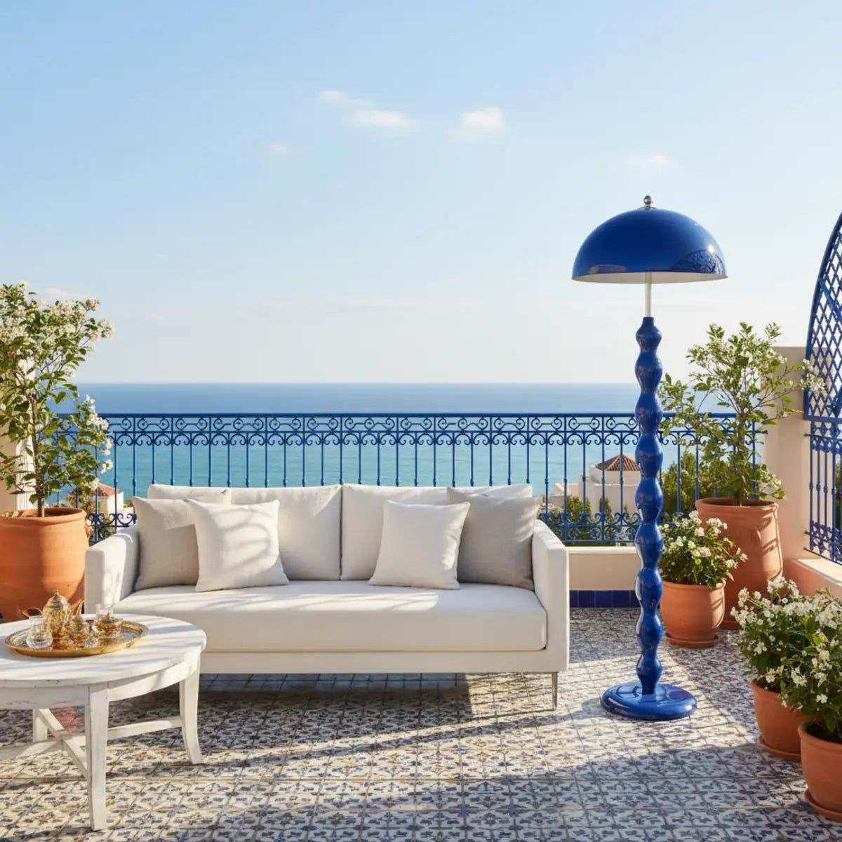 Lampadaire bleu sur une terrasse avec canapé blanc, garde-corps bleu en fer forgé et vue sur la mer