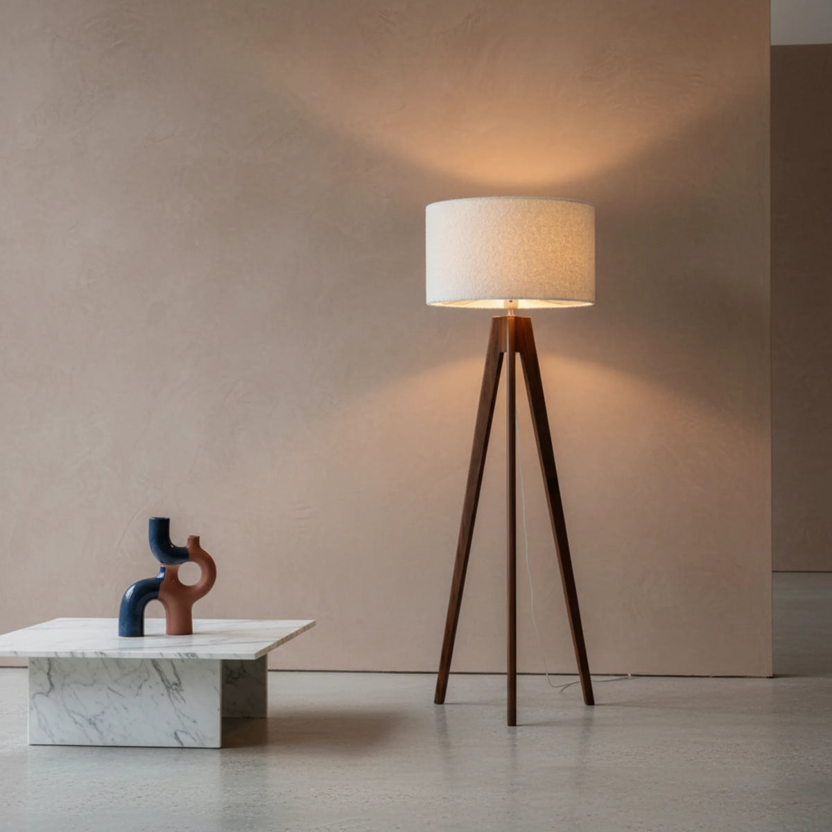 Lampadaire trépied en bois foncé allumé contre un mur beige, à côté d'une table basse en marbre blanc