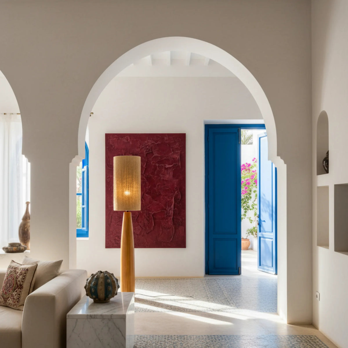 Lampadaire en bois et jute allumé sous une arche blanche, près d'une porte bleue et d'un tableau rouge