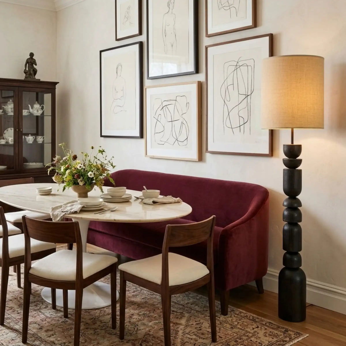 Lampadaire colonne dans un coin repas avec banquette en velours bordeaux, table ronde blanche et mur de dessins encadrés