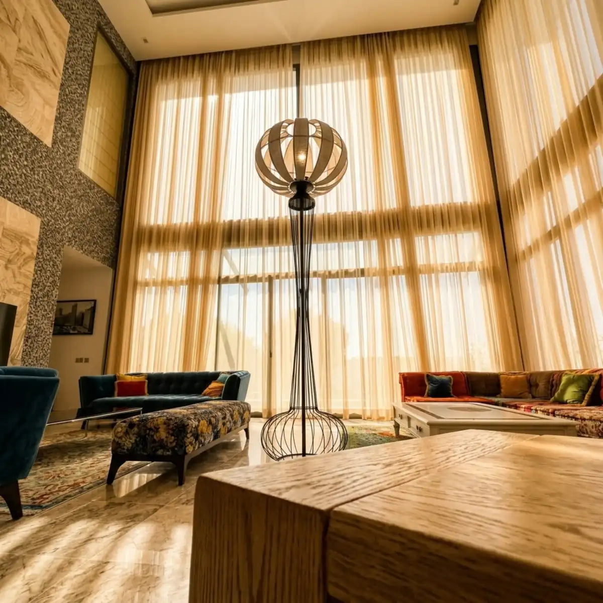 Lampadaire design en métal noir dans un grand salon aux hauts voilages, près d'une table en bois massif et de canapés colorés
