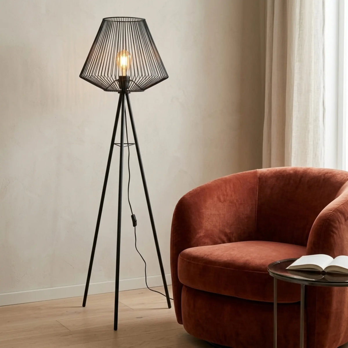 Lampadaire filaire noir allumé près d'un fauteuil en velours rouille et d'une petite table avec livre ouvert