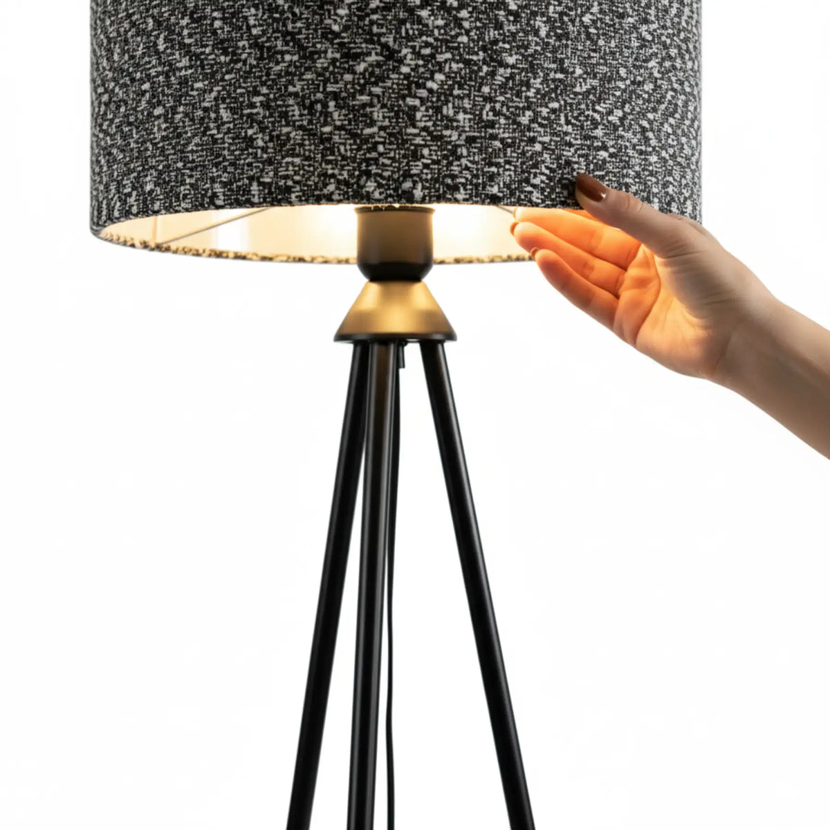 Main touchant l'abat-jour chiné du lampadaire, détail de la jonction dorée et du trépied noir sur fond blanc