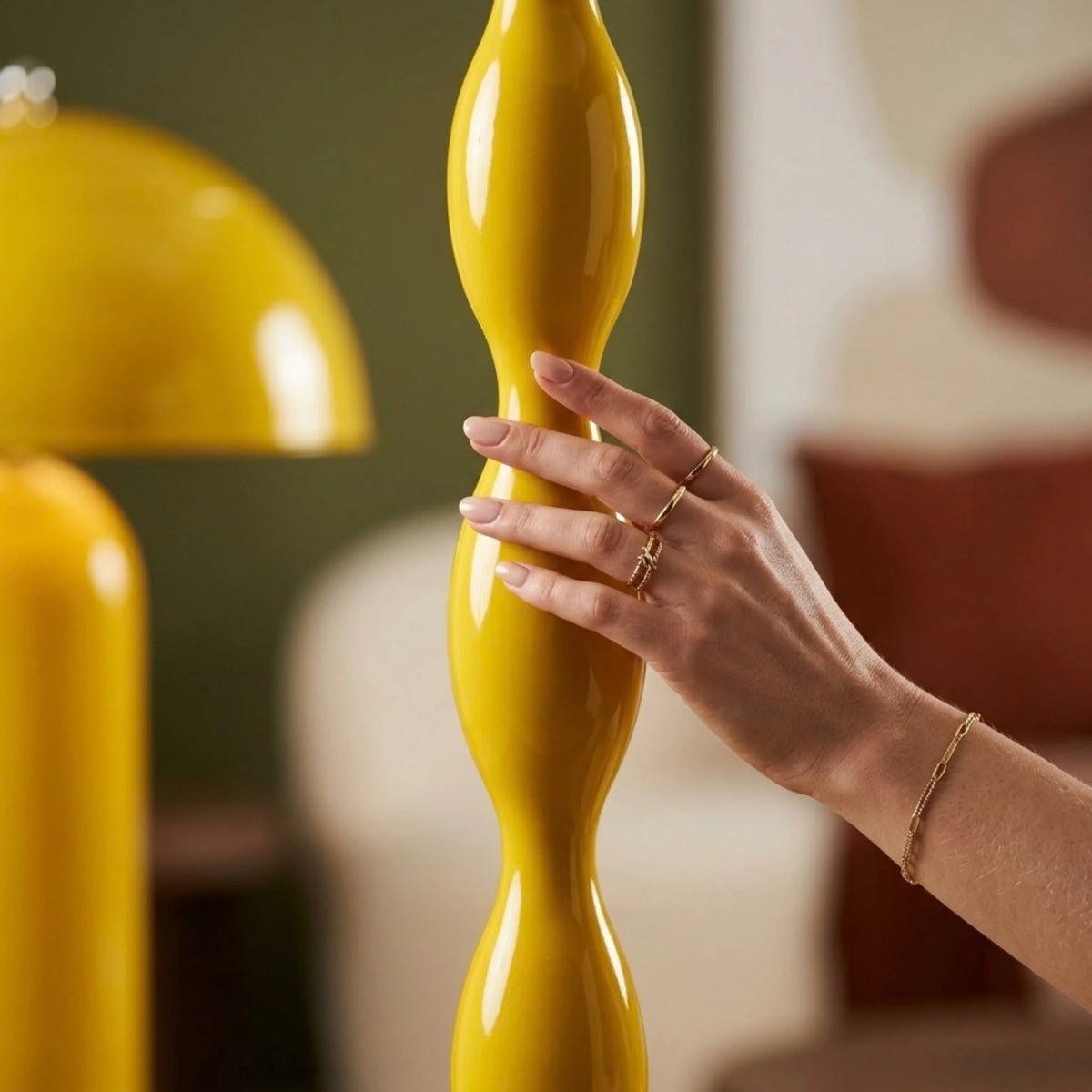 Main portant des bagues et un bracelet dorés, posée sur le pied ondulé jaune brillant du lampadaire en gros plan.