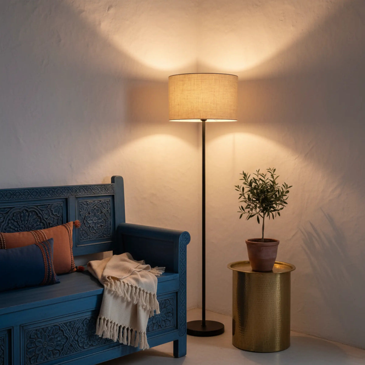 Lampadaire allumé entre un banc sculpté bleu avec coussins et un petit olivier posé sur une table d'appoint dorée