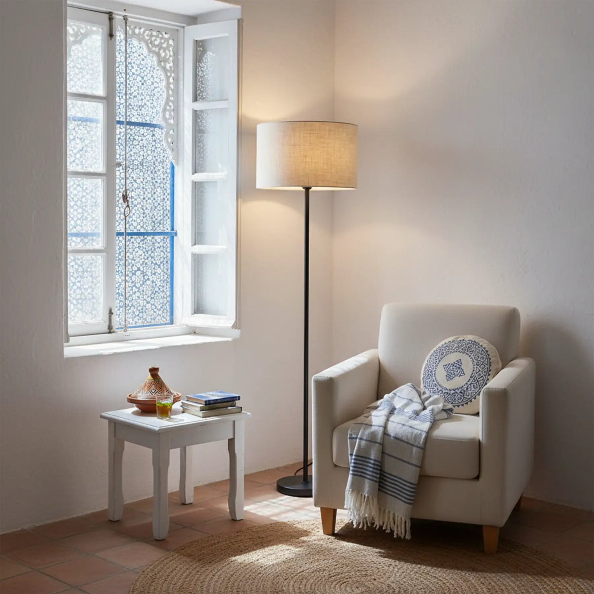 Lampadaire pied noir et abat-jour beige dans un angle près d'une fenêtre à volets blancs, fauteuil blanc et petite table