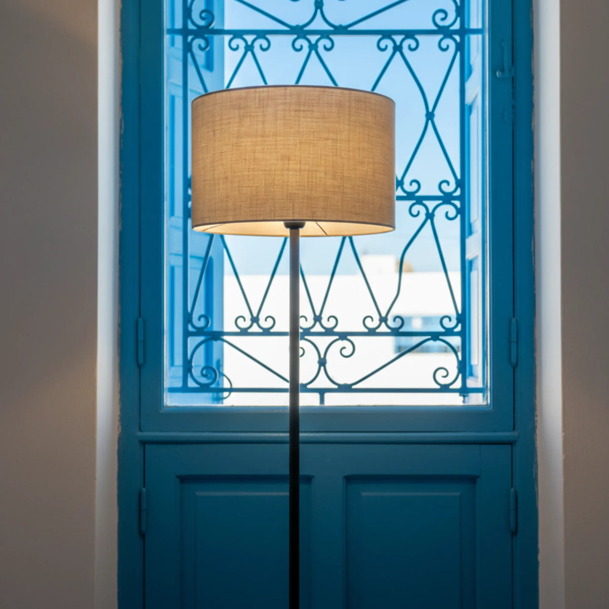 Abat-jour beige du lampadaire devant une porte-fenêtre bleue à grille en fer forgé, style méditerranéen