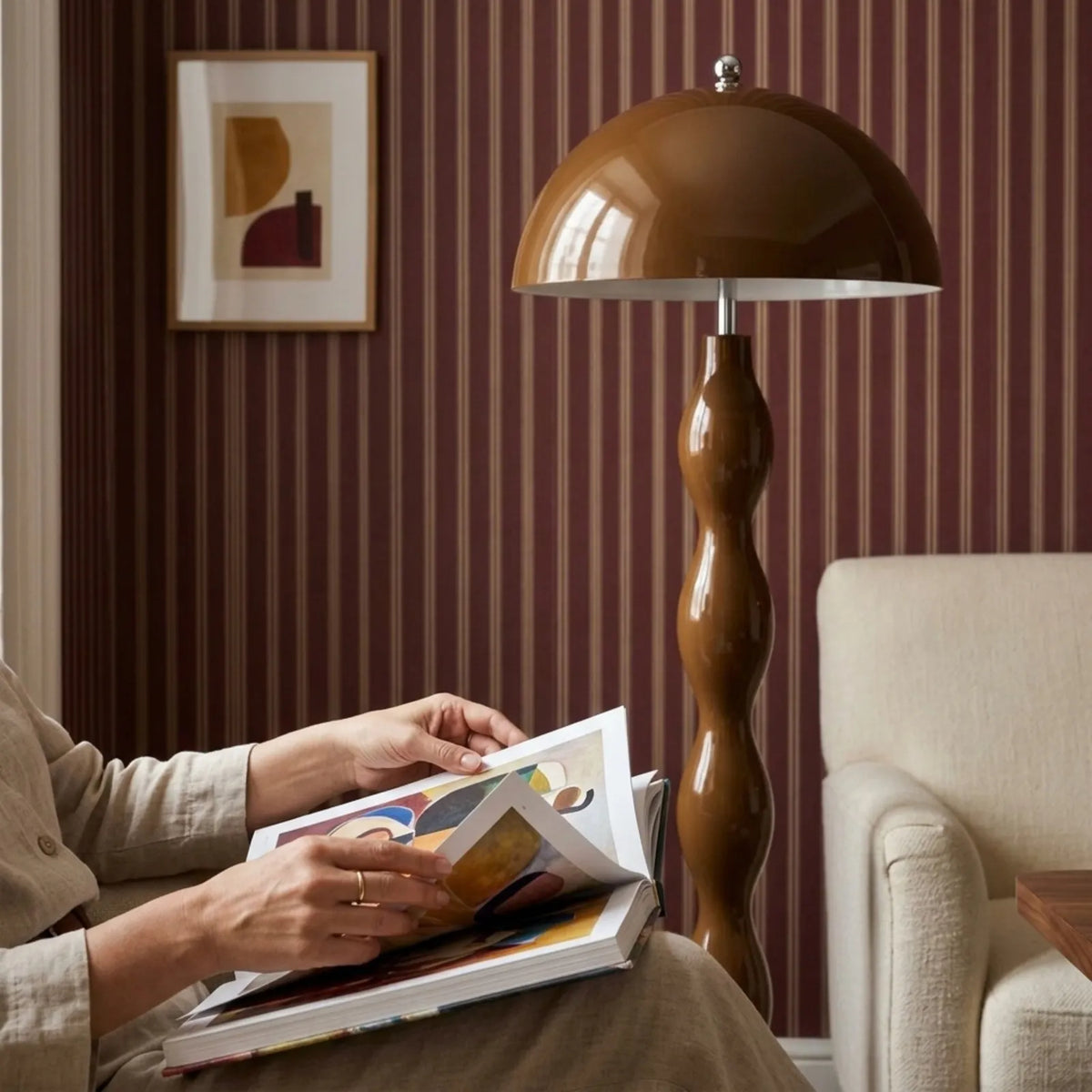 Lampadaire marron près d'un fauteuil crème où une personne lit un livre, devant un papier peint rayé bordeaux