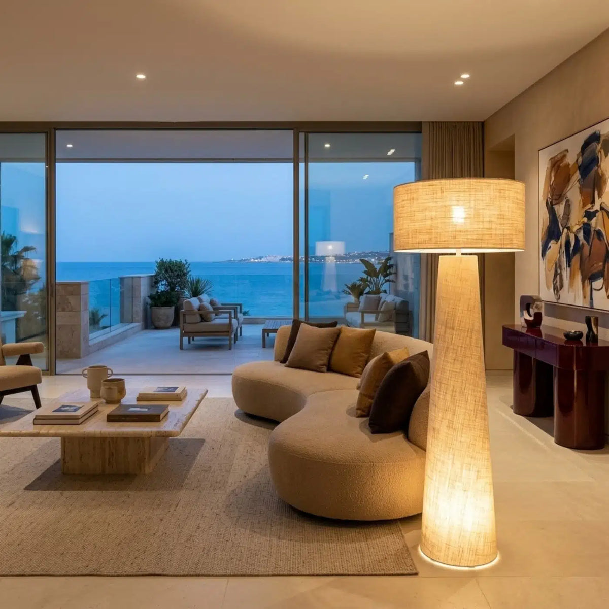 Lampadaire en lin allumé près d'un canapé courbe beige, dans un grand salon ouvert sur la mer