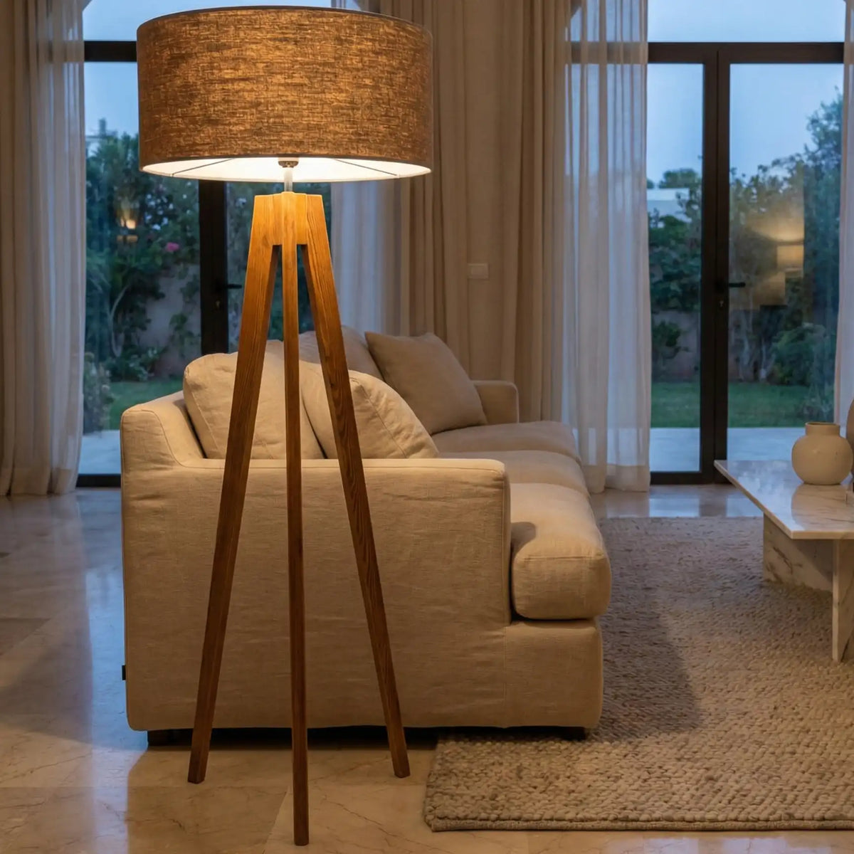 Lampadaire scandinave trépied allumé le soir devant un canapé en lin beige, baie vitrée sur le jardin