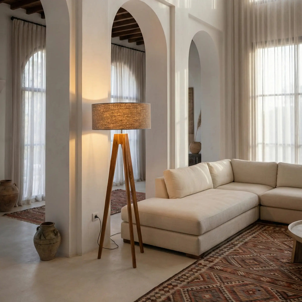 Lampadaire scandinave trépied allumé dans un salon à arches, à côté d'un canapé d'angle crème et d'un tapis rouge