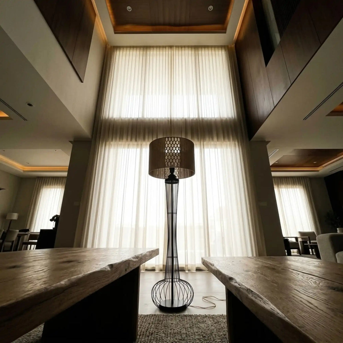 Lampadaire à abat-jour tambour en jute et macramé dans un grand salon aux hauts voilages, vu entre deux tables en bois