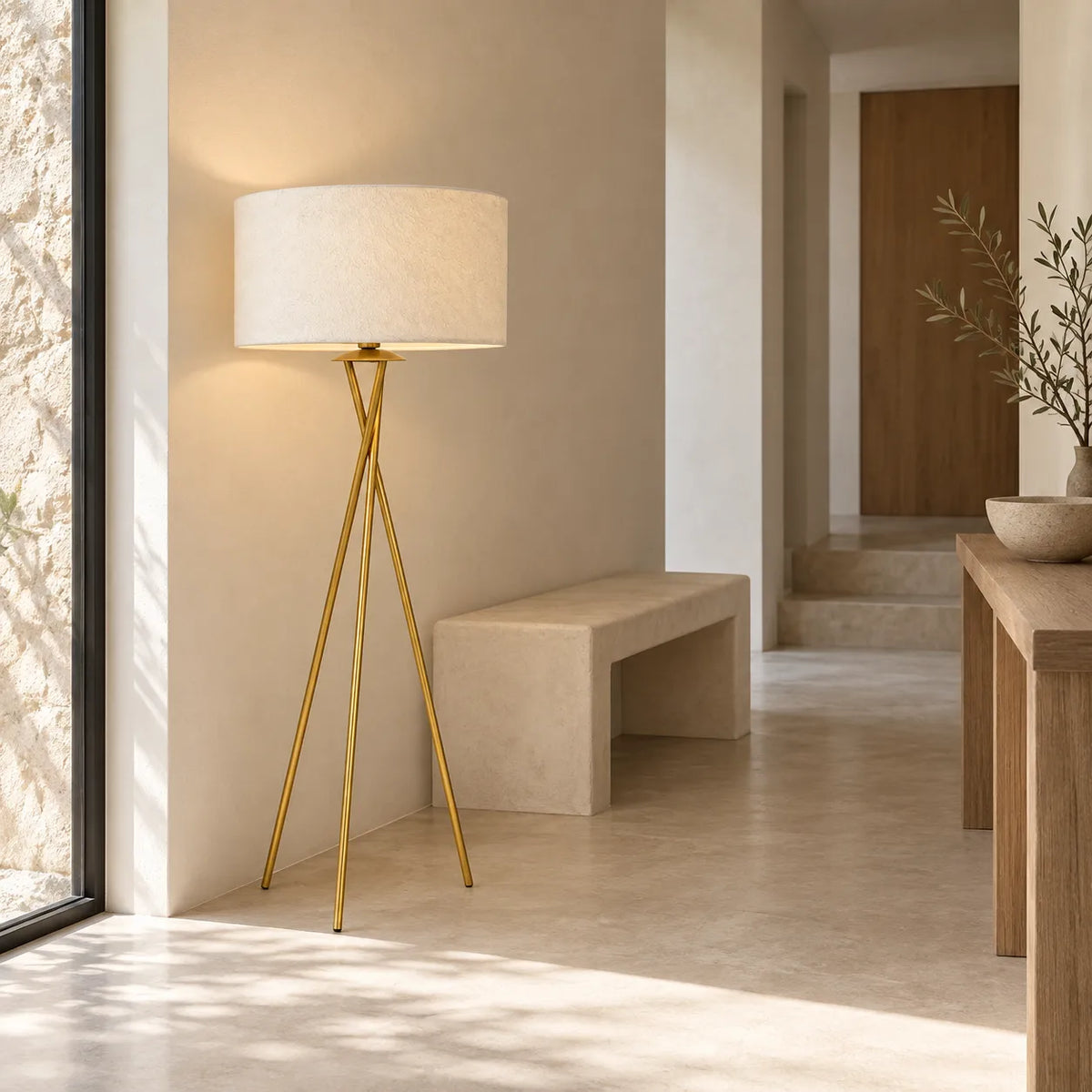 Lampadaire trépied doré allumé dans une entrée lumineuse, à côté d'un banc en pierre et d'une console en bois