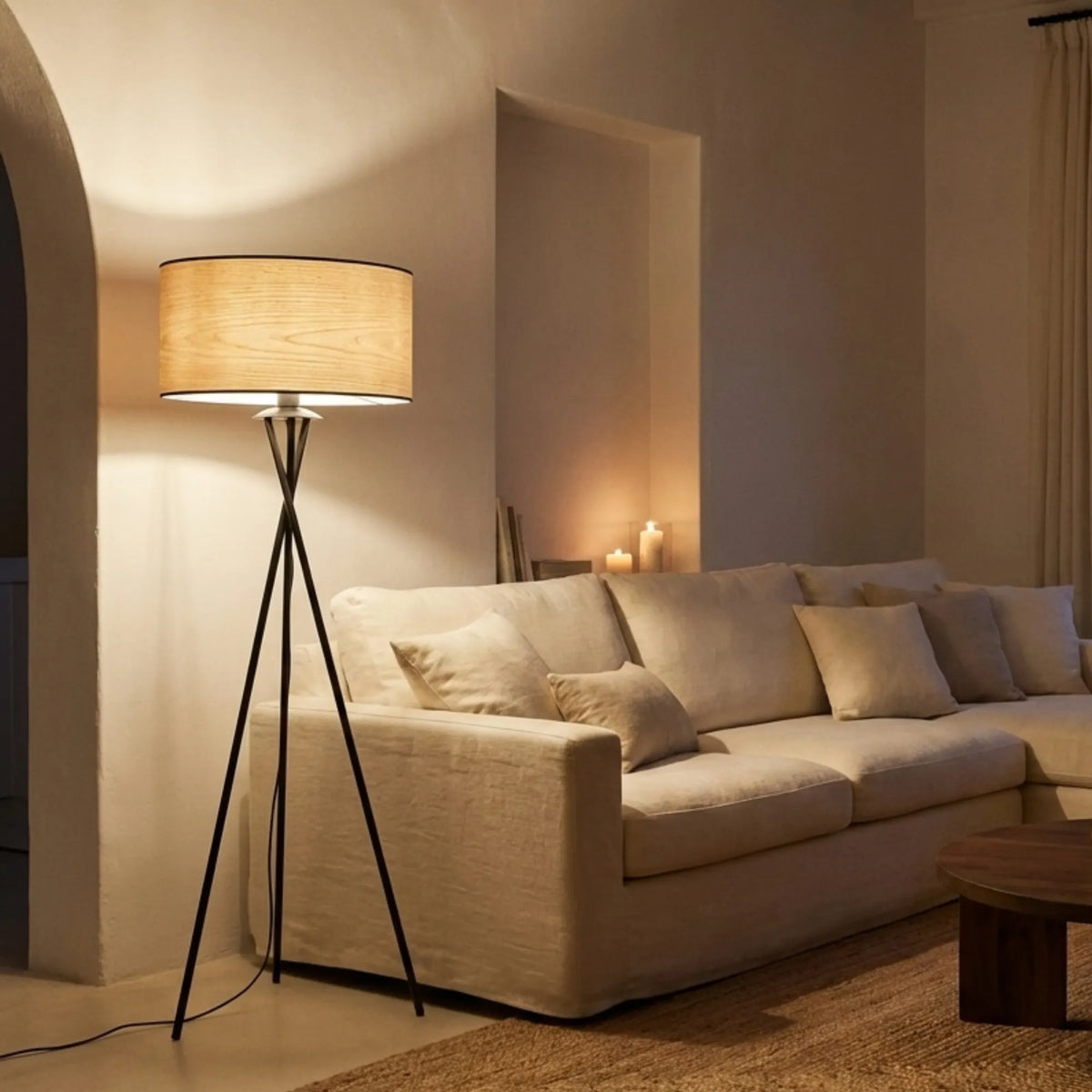 Lampadaire trépied noir allumé le soir à côté d'un canapé beige, lumière douce à travers l'abat-jour effet bois