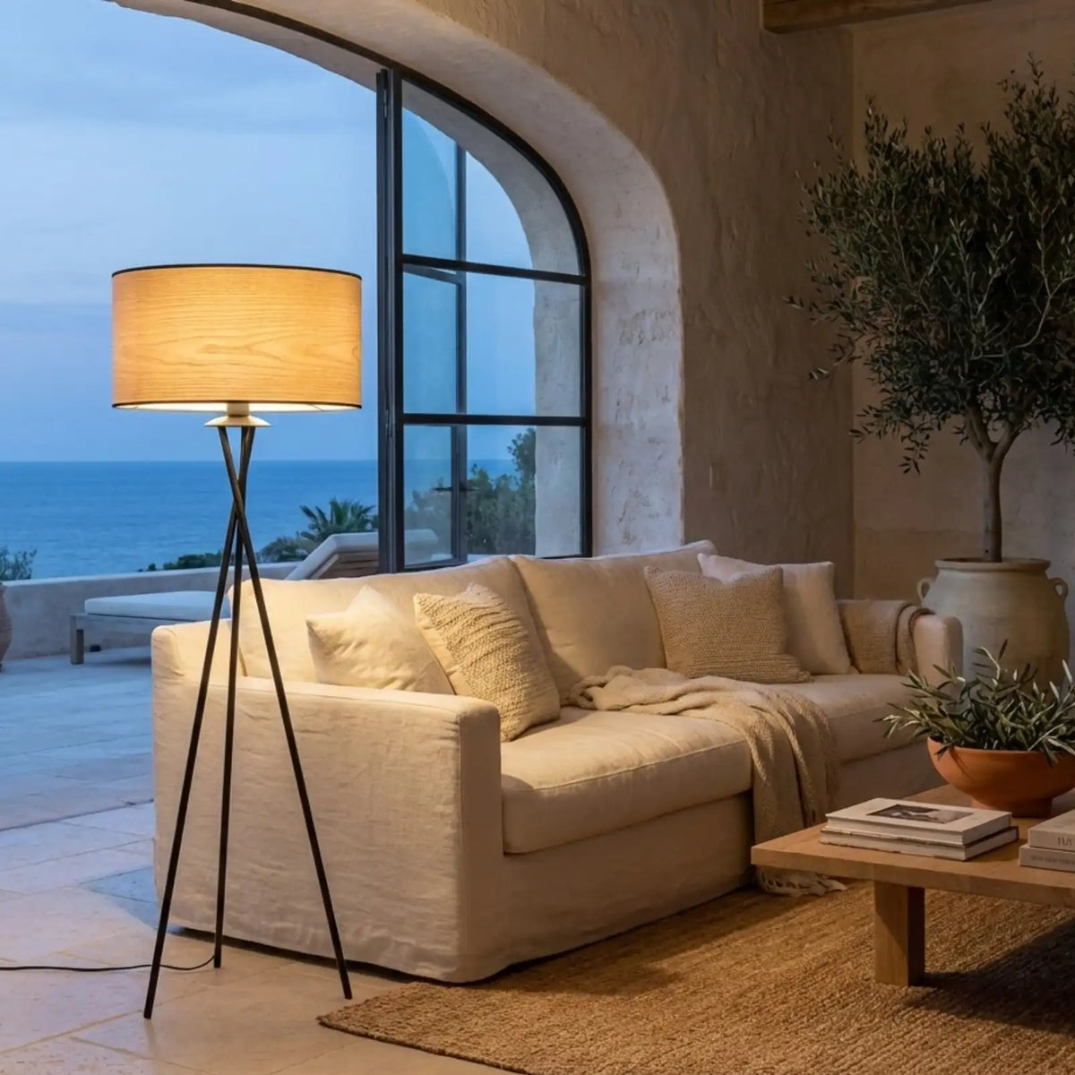 Lampadaire trépied noir allumé au crépuscule près d'un canapé en lin, d'un olivier et d'une grande baie vitrée face à la mer