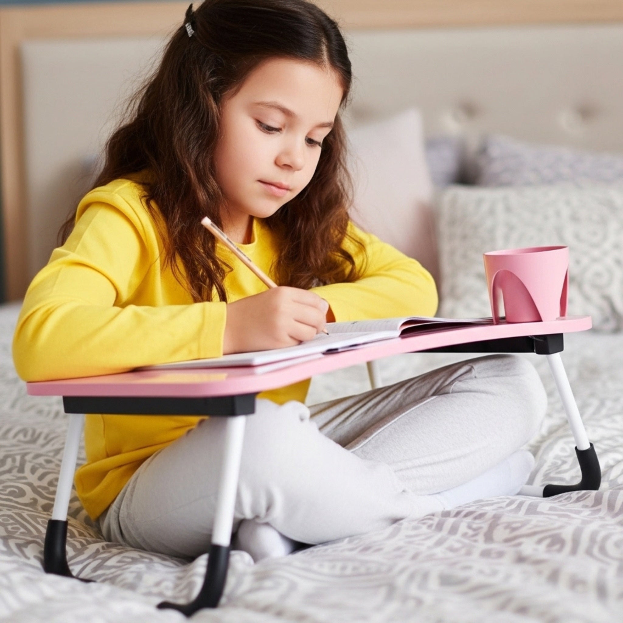 Une jeune fille fait ses devoirs sur une table de lit pliante rose, un accessoire idéal pour les enfants en Tunisie.
