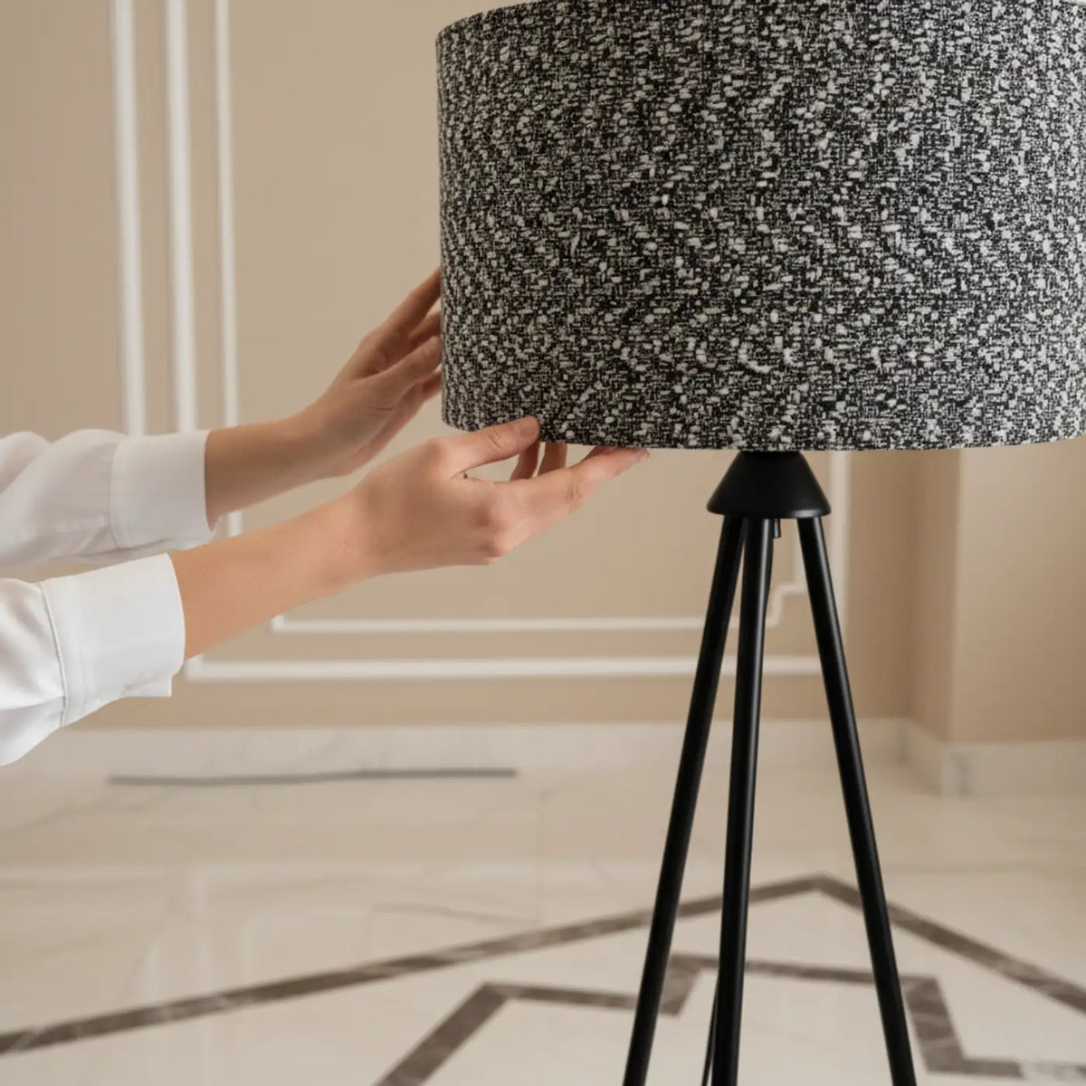 Mains posant l'abat-jour tambour en tissu chiné sur le trépied en métal noir, sol en marbre