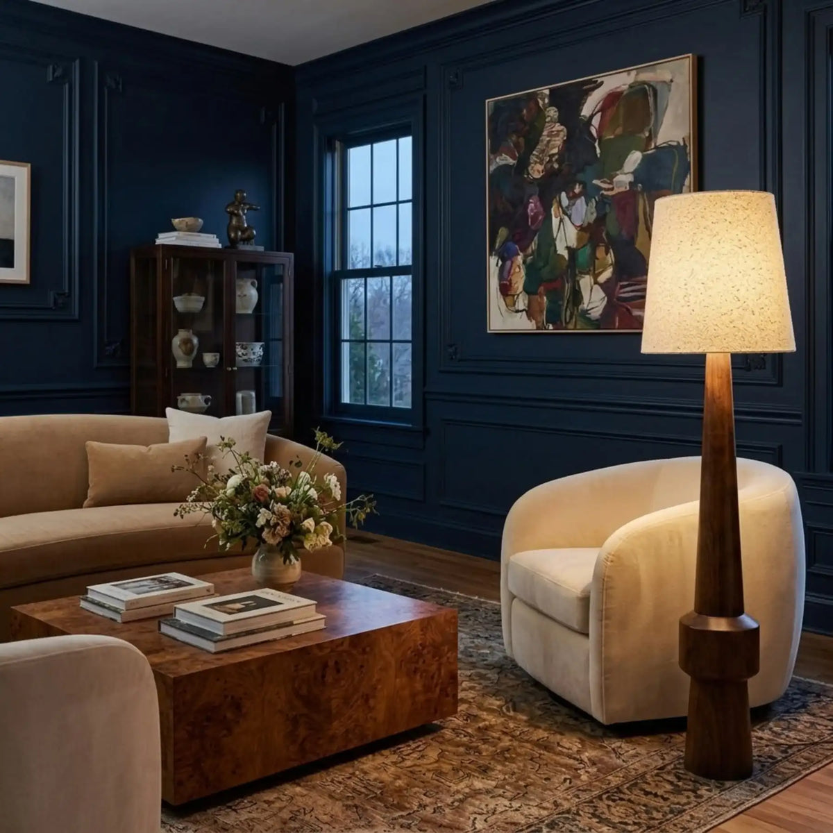 Lampadaire Atlas allumé dans un salon aux murs bleu nuit, à côté d'un fauteuil arrondi crème et d'une table basse en bois