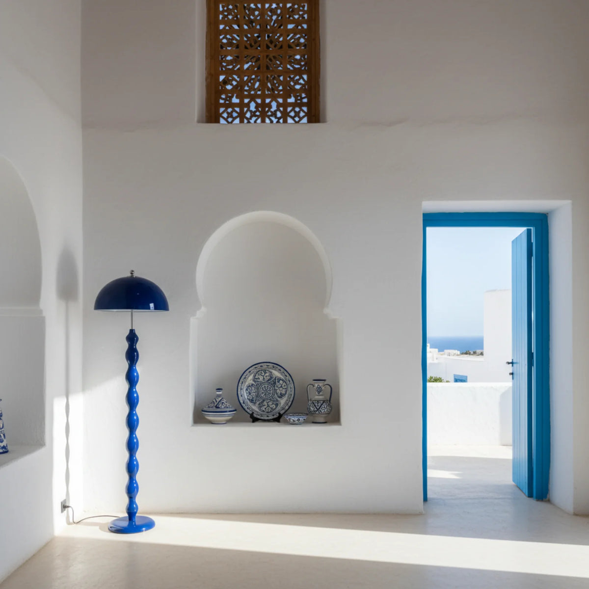 Lampadaire bleu dans une pièce blanche de style tunisien, près d'une niche en arc avec céramiques bleues