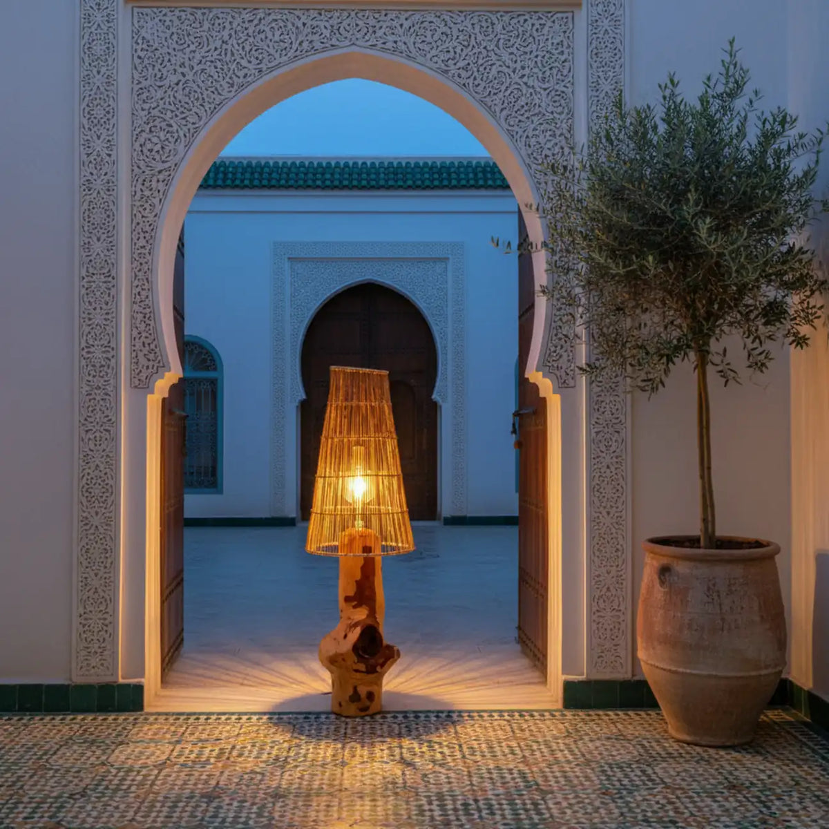 Lampadaire en bois allumé sous une porte en arc sculpté, à côté d'un olivier en pot