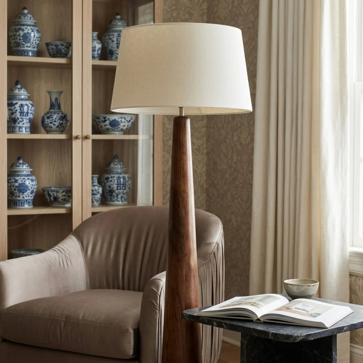 Coin lecture avec lampadaire en bois tourné, fauteuil taupe, livre ouvert et vitrine de porcelaines bleues et blanches