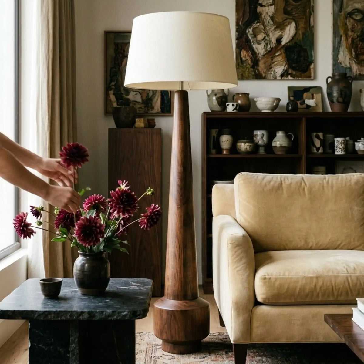 Lampadaire en bois tourné entre un fauteuil beige et une table d'appoint où une main arrange un bouquet de dahlias