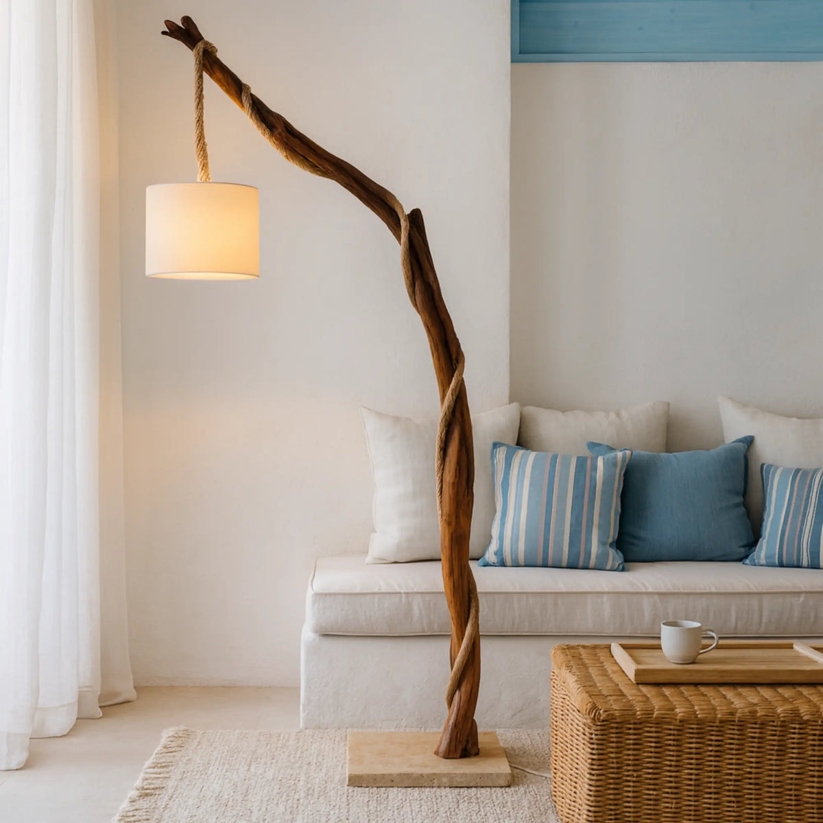 Lampadaire sur pied en bois naturel allumé à côté d'un canapé blanc aux coussins bleus rayés et d'un pouf en osier