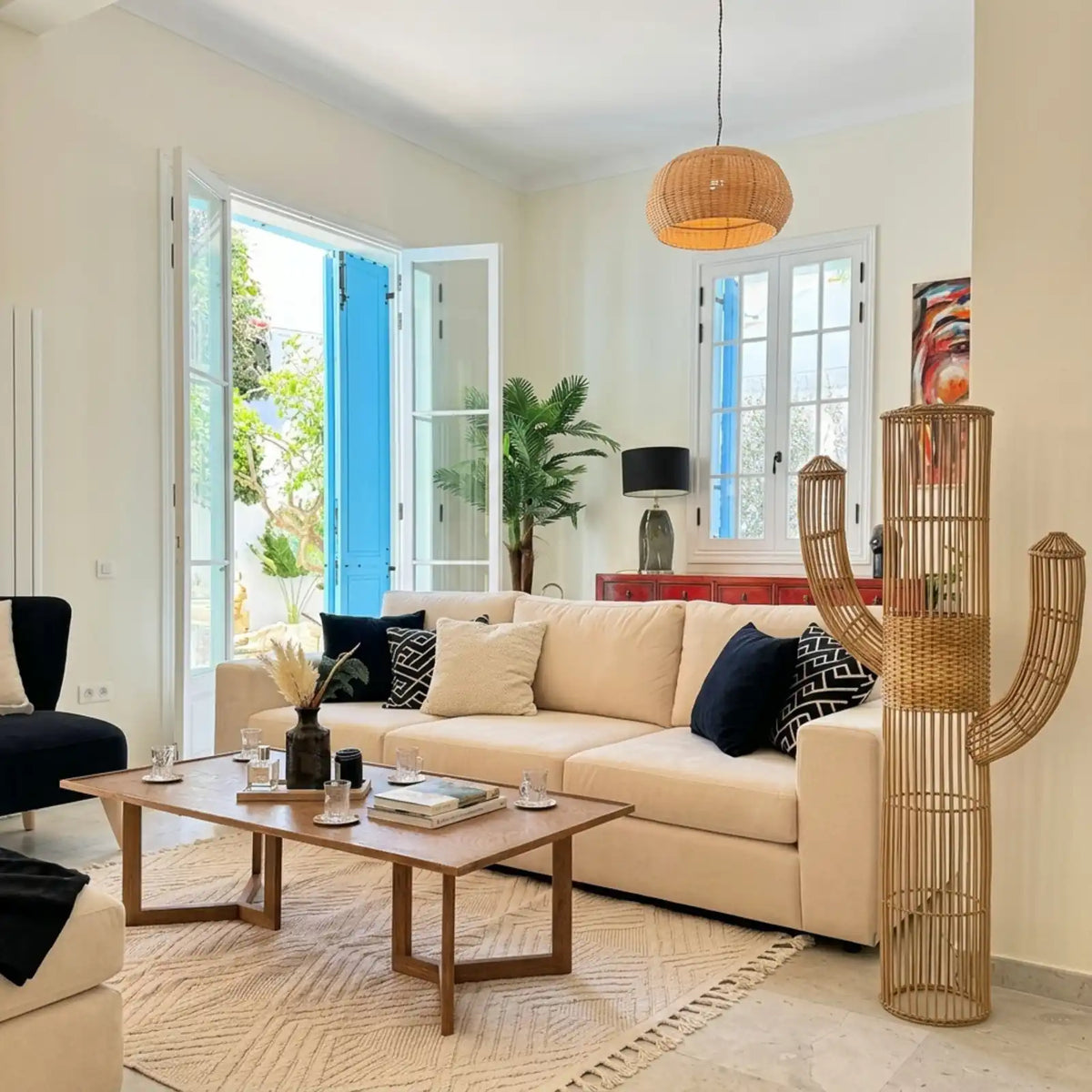 Lampadaire cactus et suspension en rotin dans un salon clair aux portes-fenêtres bleues, à côté d'un canapé crème