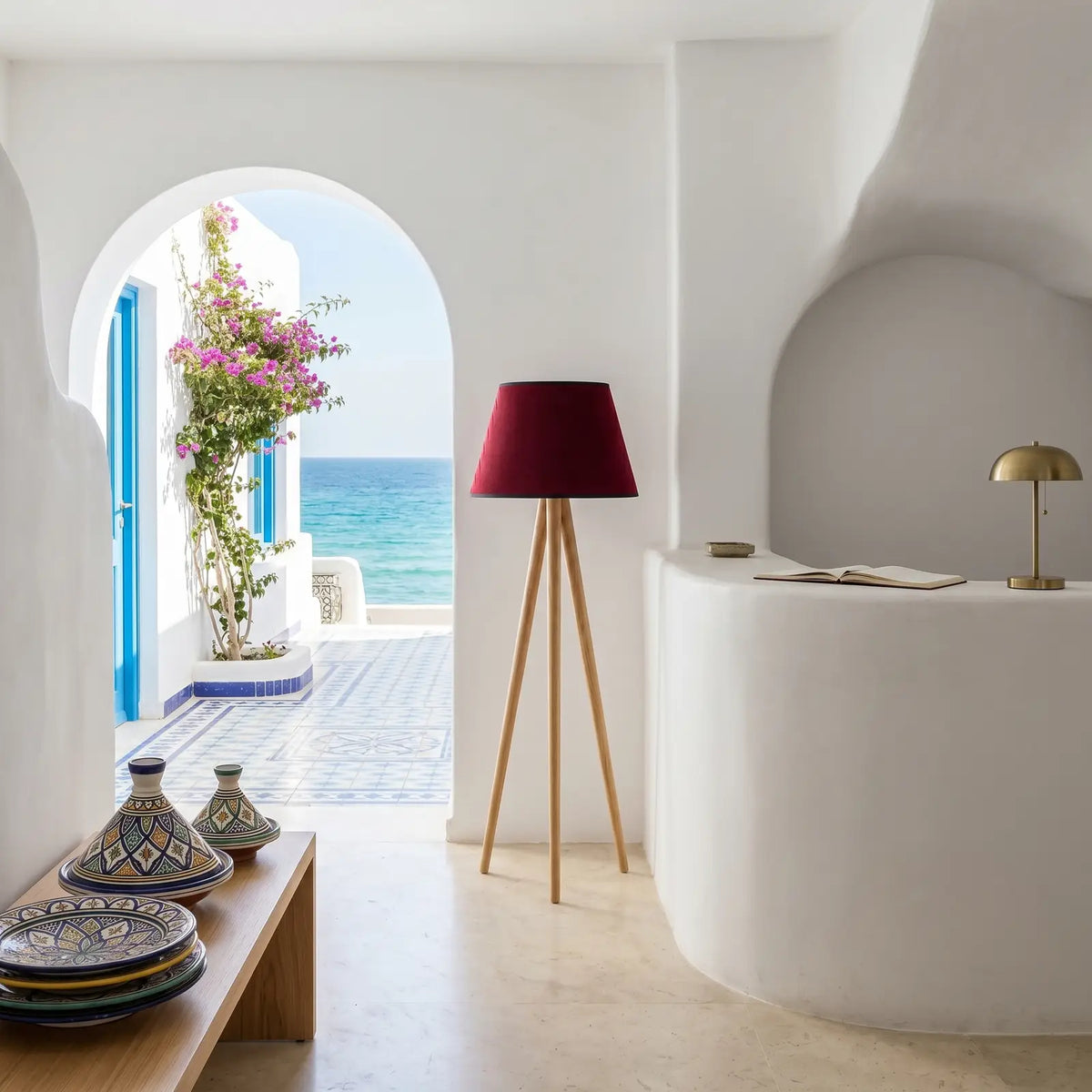 Lampadaire trépied en bois dans une entrée blanche aux murs arrondis, avec une arche ouvrant sur la mer