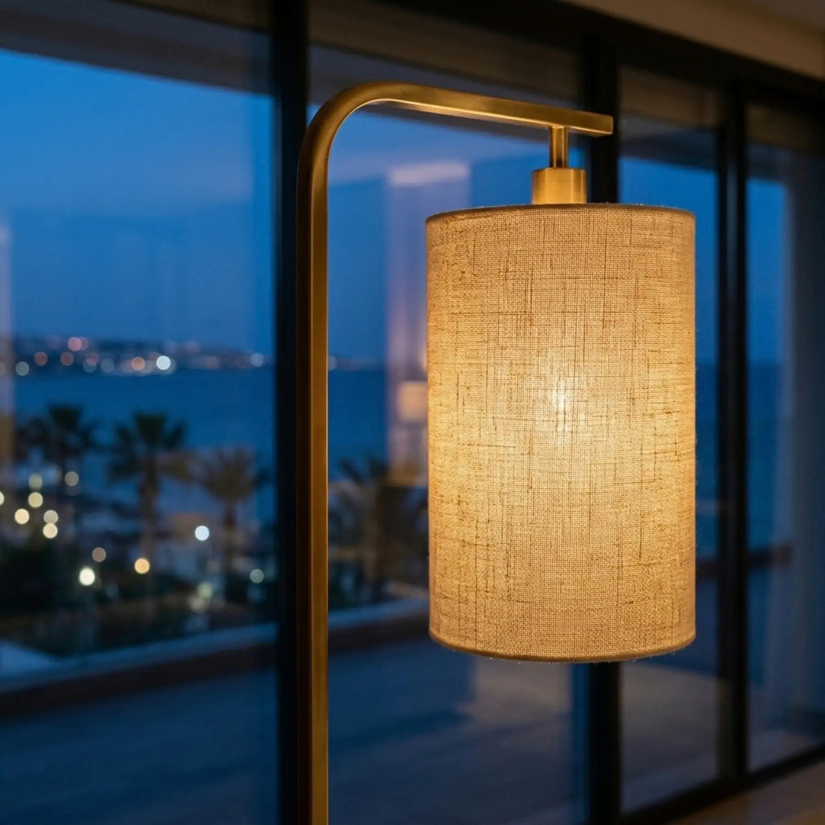 Abat-jour en lin allumé du lampadaire doré vu de près, devant une baie vitrée de nuit