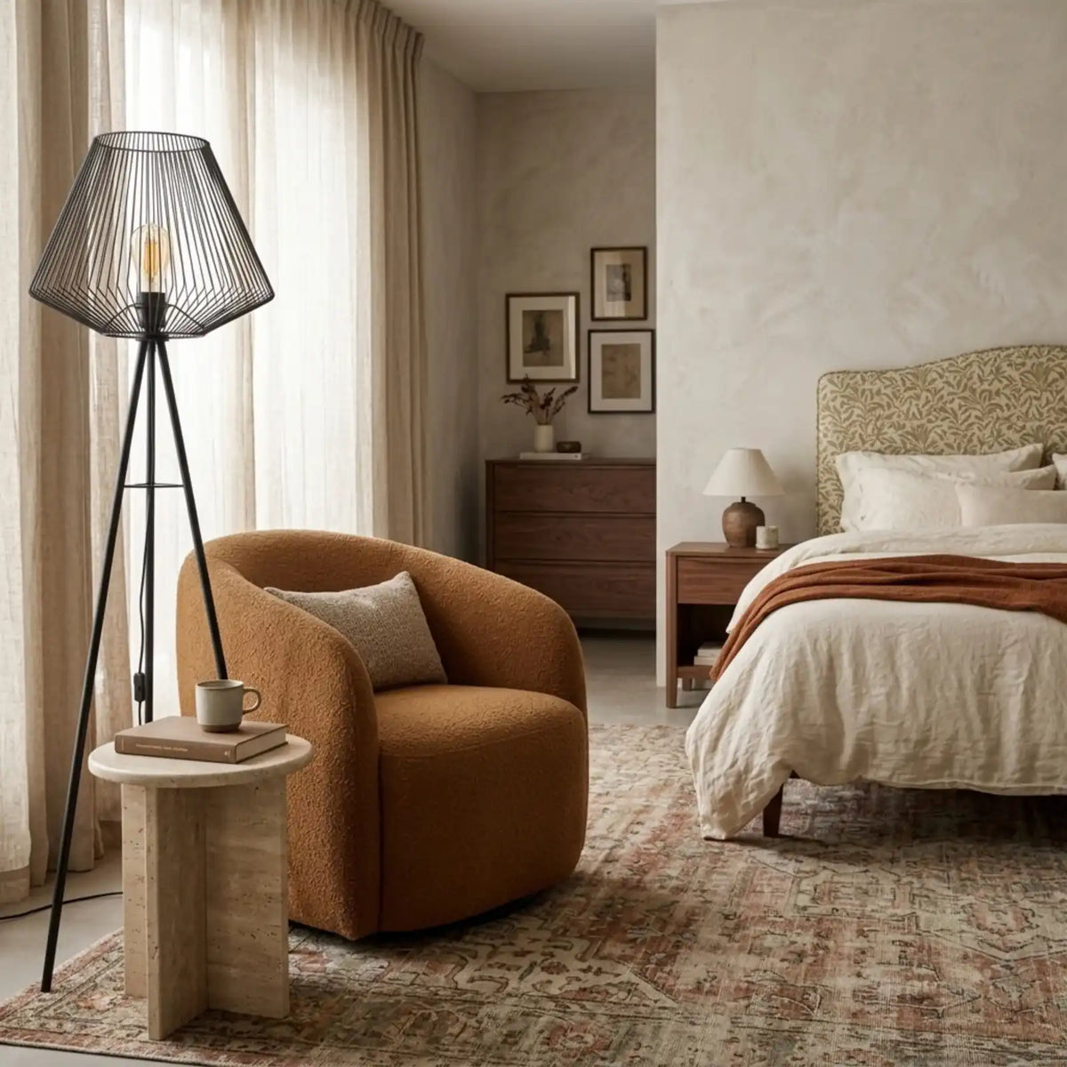 Lampadaire filaire trépied dans une chambre, à côté d'un fauteuil arrondi camel et d'une table d'appoint, lit en arrière-plan