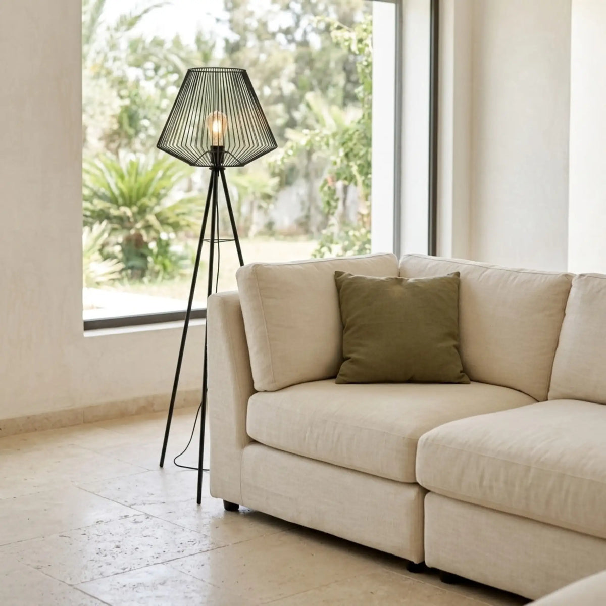 Lampadaire filaire noir derrière un canapé d'angle blanc avec coussin vert, devant une grande fenêtre sur jardin
