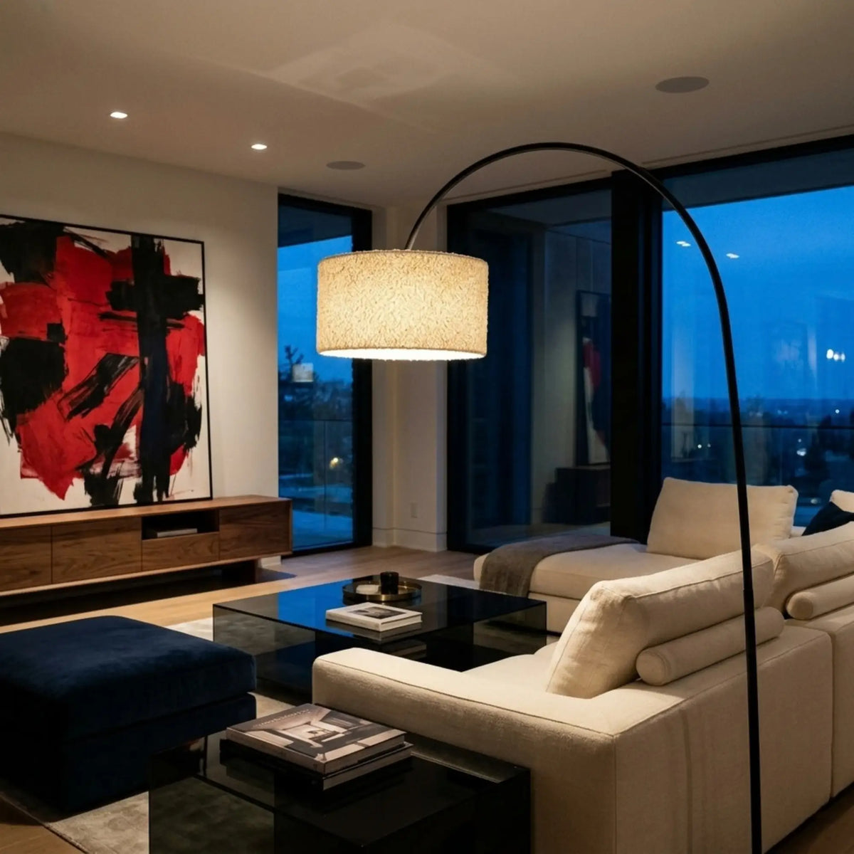 Lampadaire girafe allumé le soir au-dessus d'un canapé crème, devant une baie vitrée et un tableau rouge et noir