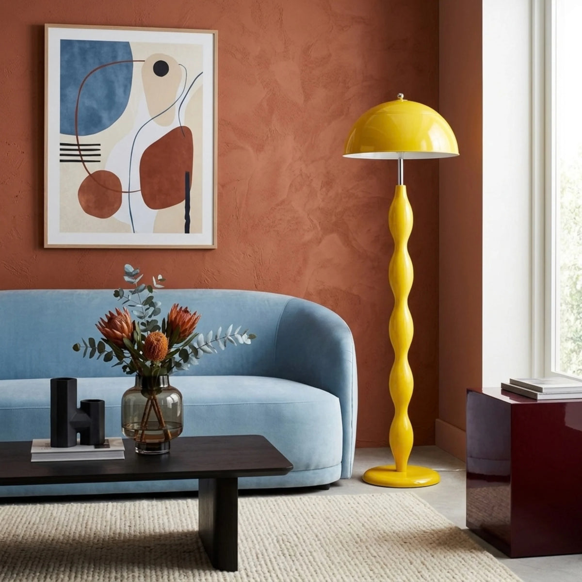 Lampadaire jaune à pied ondulé à côté d’un canapé bleu clair, devant un mur terracotta et une affiche abstraite.