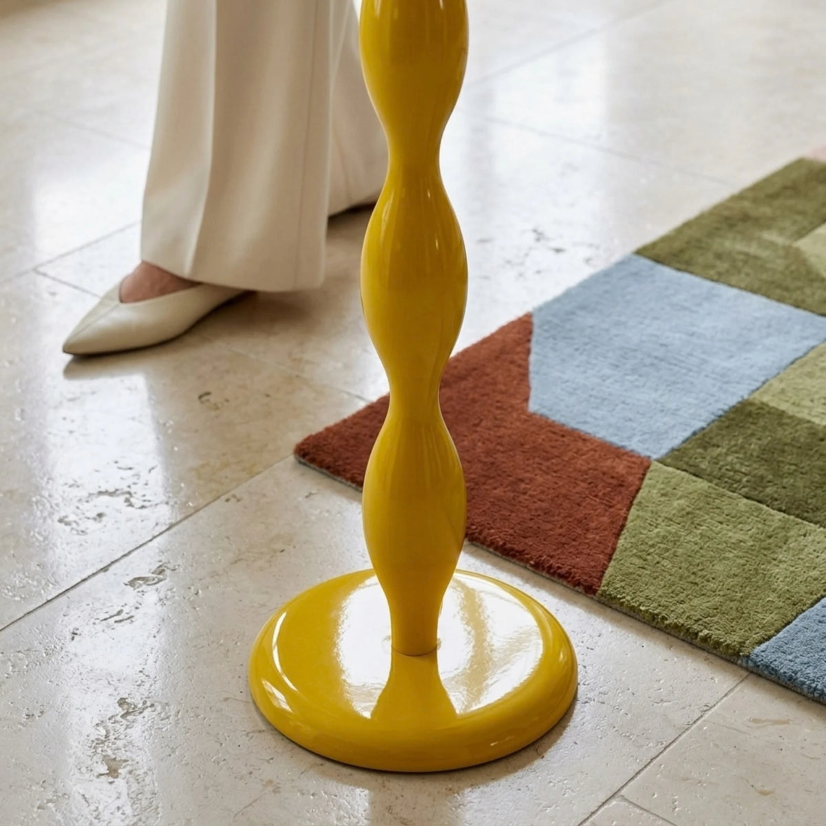 Détail du socle rond et du bas du pied ondulé jaune brillant du lampadaire, près d’un tapis géométrique coloré.