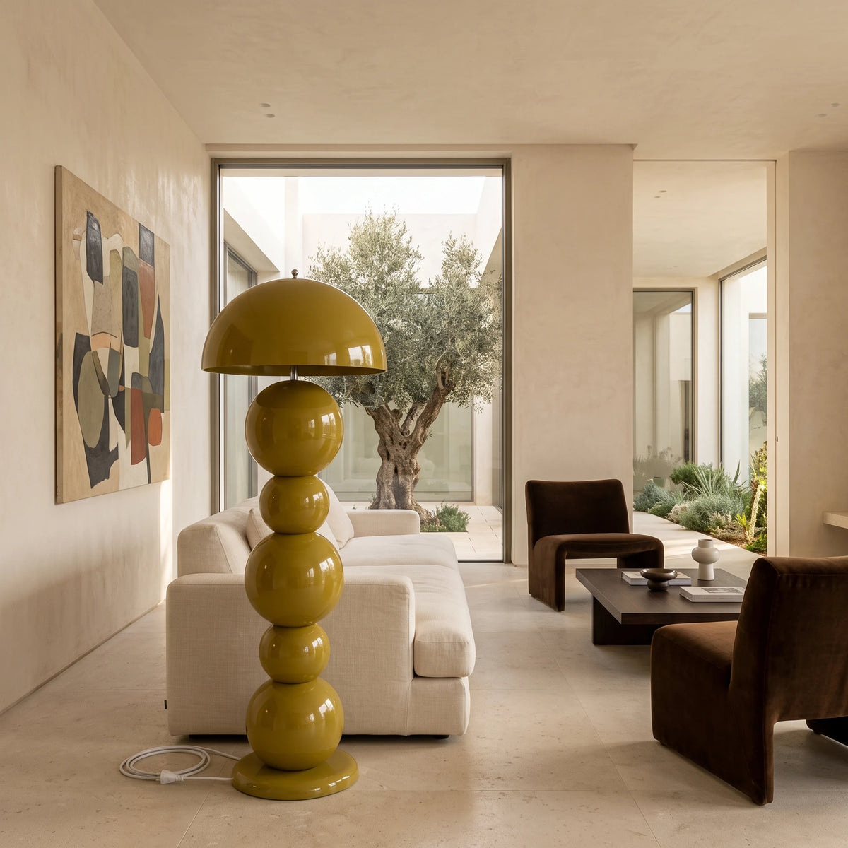 Lampadaire vert olive devant une baie vitrée ouverte sur un olivier, avec canapé blanc et fauteuils en cuir
