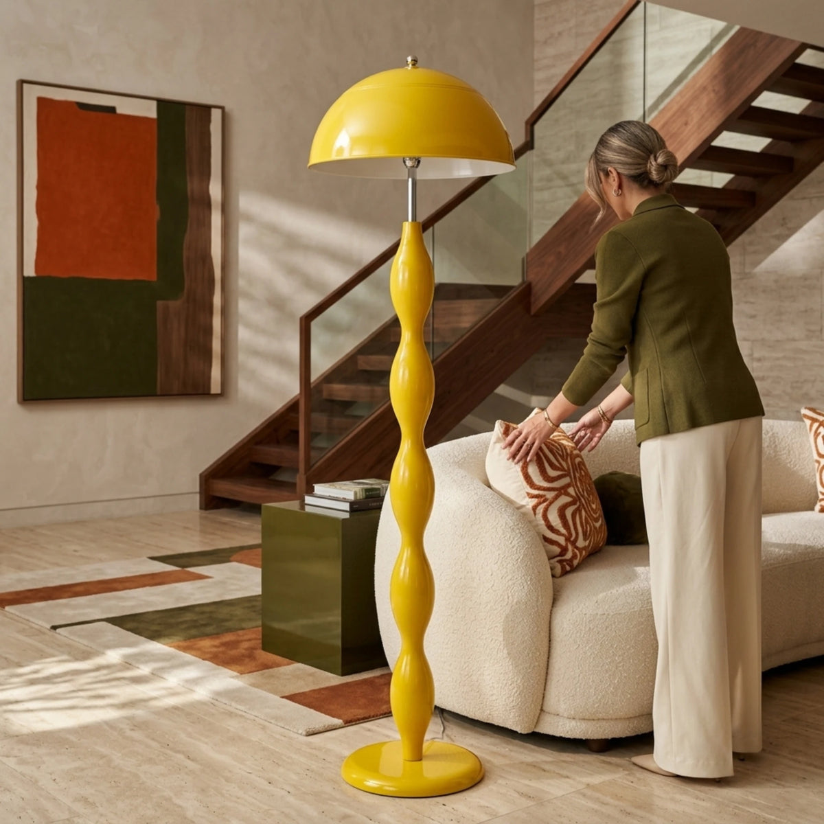 Lampadaire jaune à pied ondulé près d’un fauteuil crème, avec une femme ajustant un coussin devant un escalier en bois.