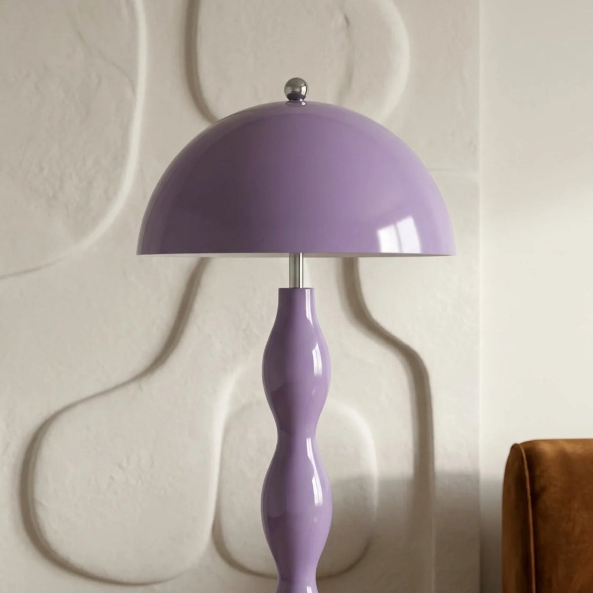 Gros plan sur l'abat-jour dôme lilas et le haut du pied ondulé, devant un mur sculpté en relief