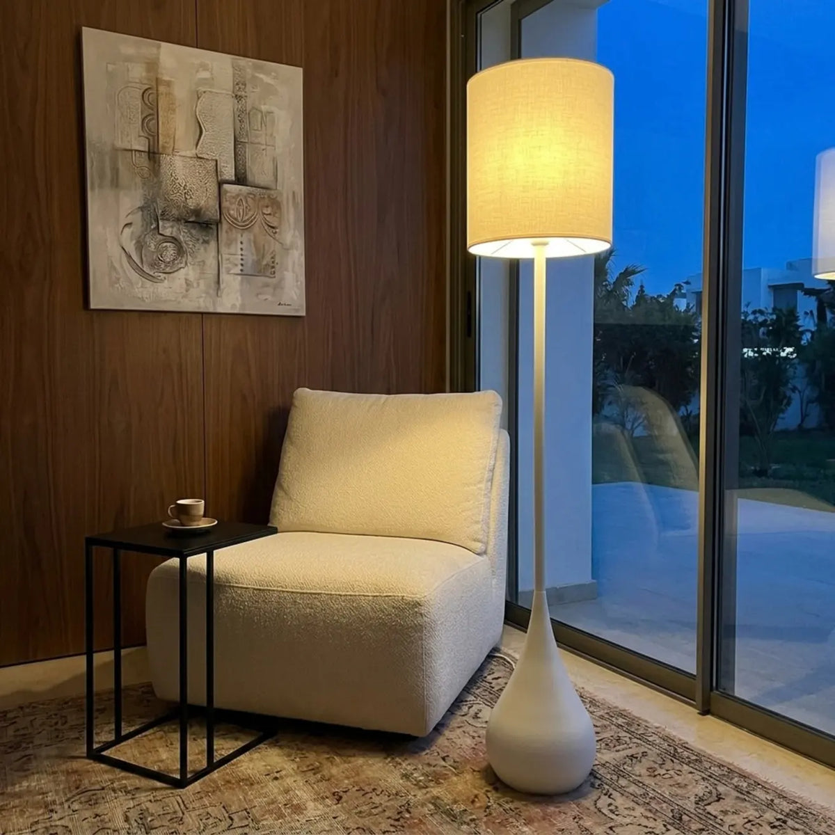 Lampadaire Goutte allumé dans un coin lecture, près d'un fauteuil crème et d'une baie vitrée au crépuscule