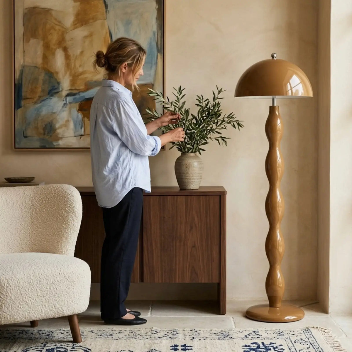 Lampadaire moderne marron à côté d'une console en noyer où une femme arrange un vase d'olivier