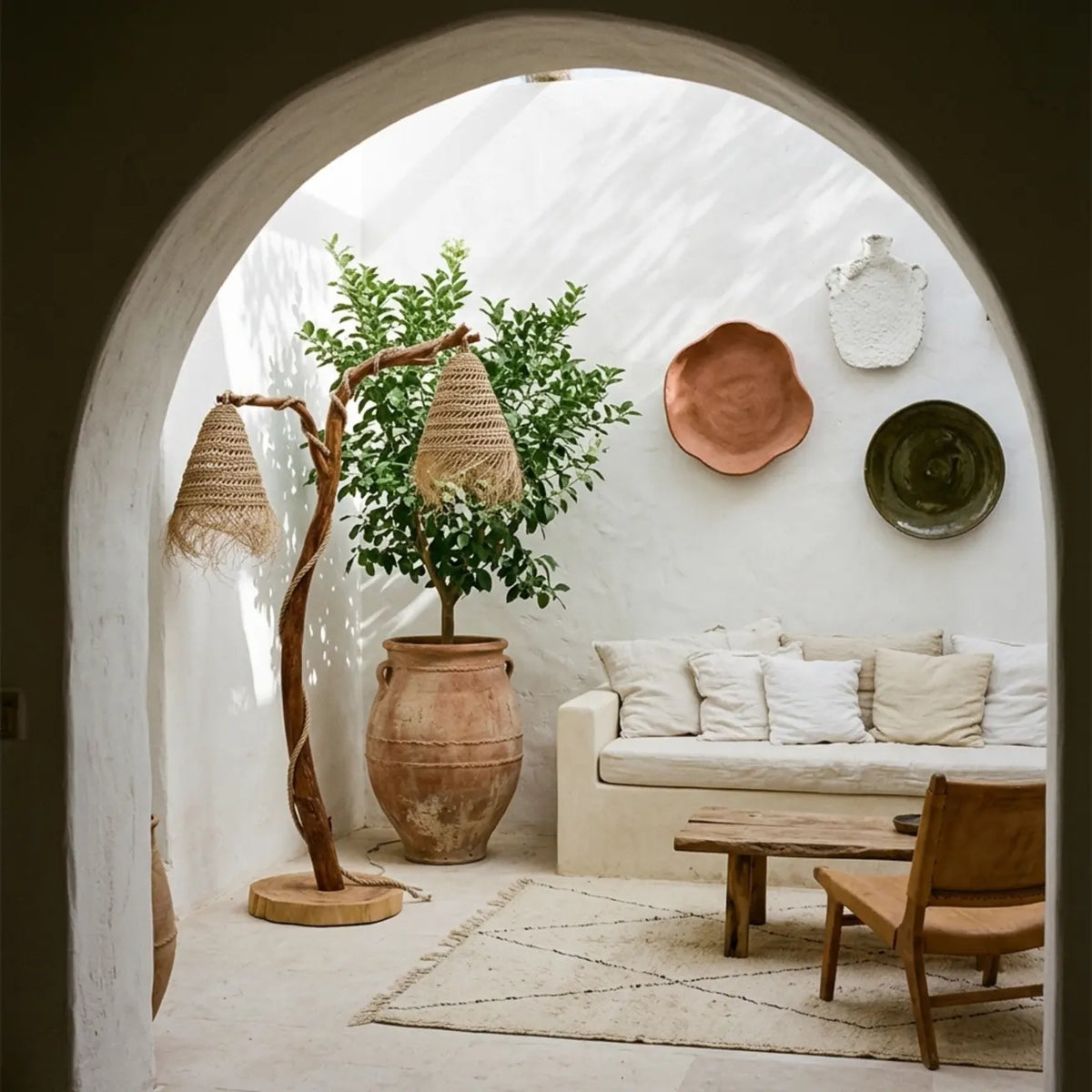 Lampadaire bohème en bois flotté vu sous une arche, dans un salon blanc avec banquette, arbre en pot et assiettes murales