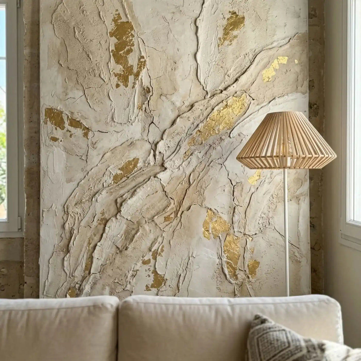 Lampadaire à abat-jour en lattes devant un tableau texturé blanc et feuille d'or, au-dessus d'un canapé crème