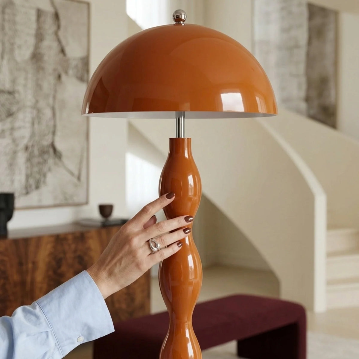 Gros plan sur l'abat-jour champignon orange laqué et le pied ondulé du lampadaire, effleuré par une main