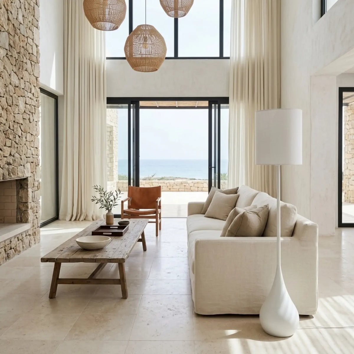Lampadaire blanc dans un grand séjour avec vue sur la mer, suspensions en rotin, mur en pierre et canapé crème