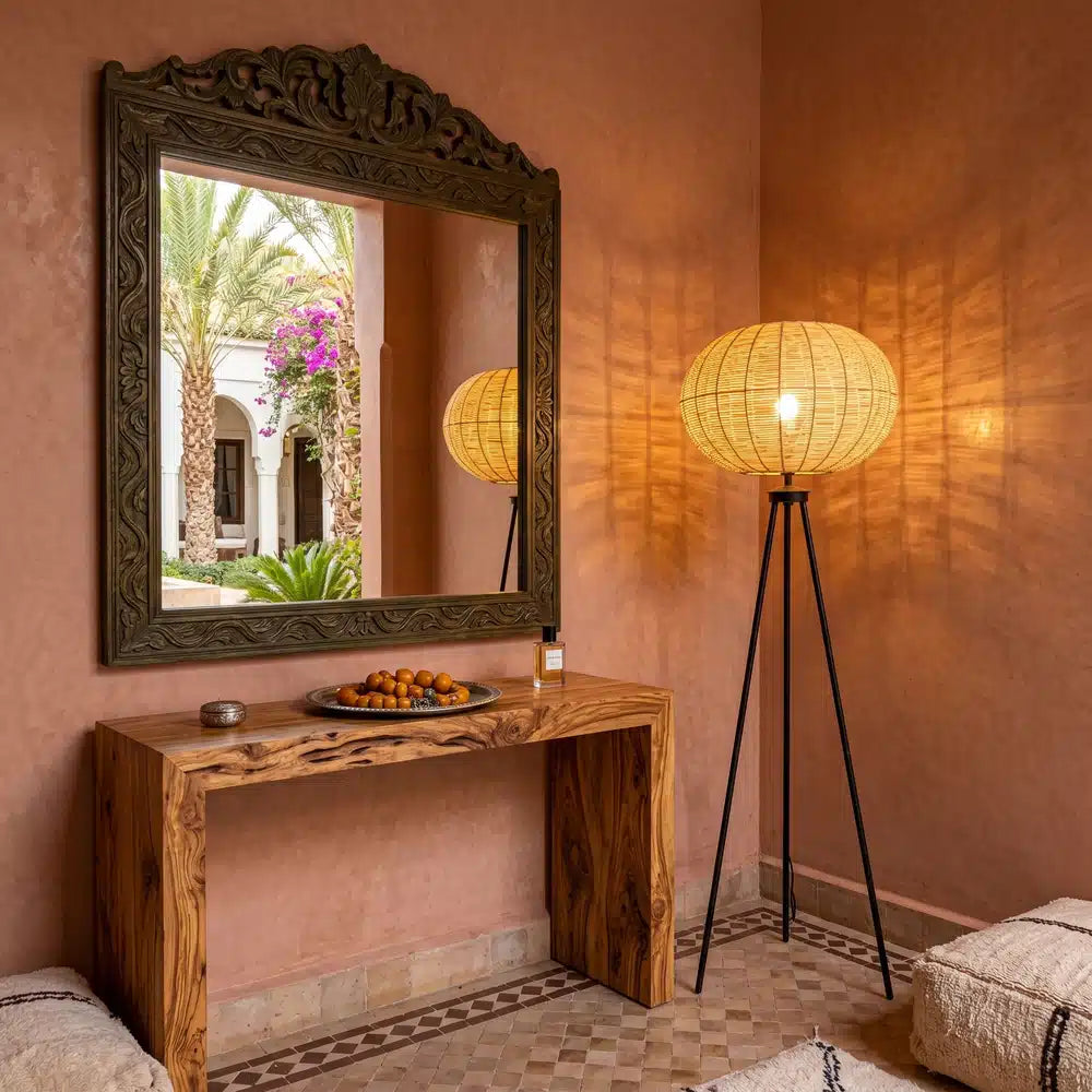 Lampadaire en rotin allumé contre un mur terracotta, à côté d'une console en bois et d'un miroir sculpté