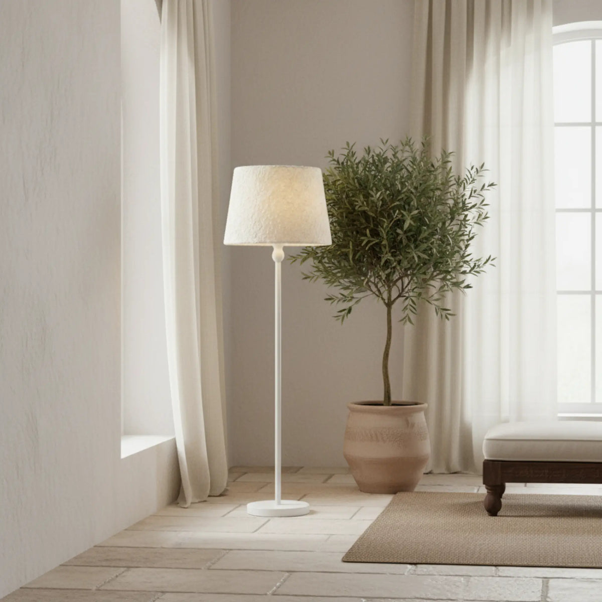 Lampadaire blanc à côté d'un olivier en pot en terre cuite, devant des voilages clairs et un banc en bois